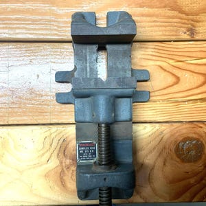 May include: A heavy-duty, dark gray Ridgid Simplex vise, model No. 31/2 D.P., with a screw mechanism for clamping. The vise is made of cast iron and features a rectangular design with a textured surface. The vise is designed for holding objects securely.