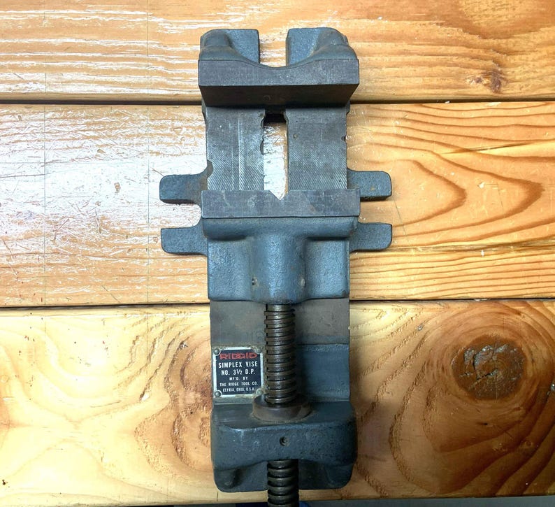 May include: A heavy-duty, dark gray Ridgid Simplex vise, model No. 31/2 D.P., with a screw mechanism for clamping. The vise is made of cast iron and features a rectangular design with a textured surface. The vise is designed for holding objects securely.