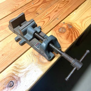 May include: A gray metal vise with a screw mechanism and a handle. The vise is resting on a wooden surface with visible wood grain. The vise has a rectangular base and a jaw for holding objects.