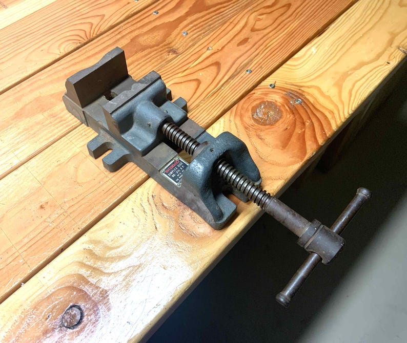 May include: A gray metal vise with a screw mechanism and a handle. The vise is resting on a wooden surface with visible wood grain. The vise has a rectangular base and a jaw for holding objects.