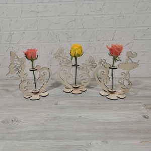 Könnte beinhalten: Drei Holz-Blumenvasen mit dekorativen Ausschnitten von Schmetterlingen und einer Ballerina. Jede Vase enthält eine einzelne Rose in Rosa- und Gelbtönen. Die Vasen stehen auf einer hellen Holzoberfläche.