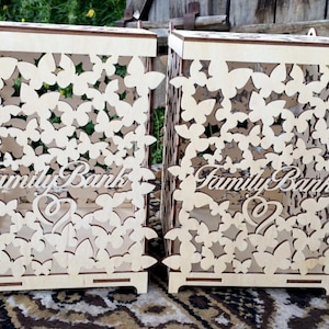 May include: Two wooden boxes with a butterfly design and the words "Family Bank" on the front. The boxes are made of light wood and have a delicate, intricate design.