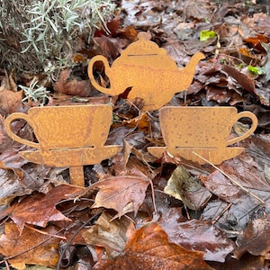 May include: Three rusty metal garden stakes shaped like a teapot and two teacups. The stakes are placed on a bed of brown leaves.