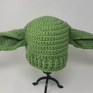 May include: A green crocheted hat with large, pointed ears. The hat is a light green color and has a ribbed brim. The ears extend outward from the sides of the hat, giving it a distinctive appearance. The hat is displayed on a black stand.