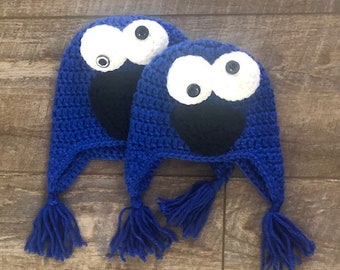 May include: Two blue crocheted hats with white and black felt appliques resembling Cookie Monster's face. Each hat has a blue tassel.