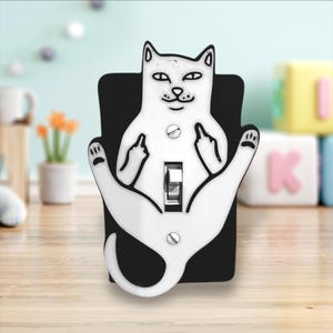 May include: A white light switch cover with a black cat design. The cat is sitting with its legs crossed and giving the middle finger with both hands. The switch cover is mounted on a black background.
