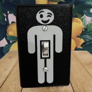 May include: A black light switch cover with a white silhouette of a person winking. The person has a simple smiley face. The switch is labeled "ON".