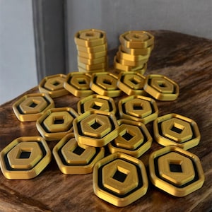 May include: A collection of gold-colored hexagonal game pieces with a black square in the center. The pieces are stacked in two piles and scattered on a wooden surface.