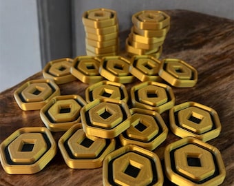 May include: A collection of gold-colored hexagonal game pieces with a black square in the center. The pieces are stacked in two piles and scattered on a wooden surface.