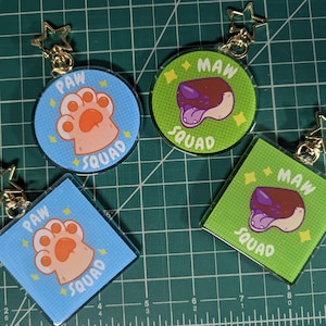May include: Four keychains with a paw print and a dog's head. The keychains are made of clear acrylic with a colorful design. The paw print keychains are blue with orange paw prints and the words "PAW SQUAD". The dog head keychains are green with a purple dog head and the words "MAW SQUAD".