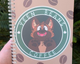 May include: A spiral-bound notebook with a light brown cover featuring a repeating pattern of dark brown coffee beans. The center displays a circular green logo with the text "YEEN BEANS COFFEE" and an illustration of a brown hyena character.