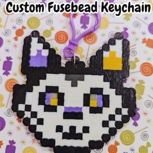 May include: A custom fusebead keychain in the shape of a cat's face. The cat is black, white, purple, and yellow. The keychain has a purple clasp. The background is a pattern of candies.
