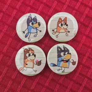 May include: Four round, white buttons with cartoon dog characters in blue, orange, and brown. Each button features a different dog character on a lined paper background. The dogs are holding a coconut drink.