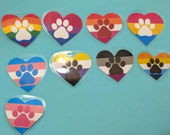May include: A collection of eight heart-shaped stickers, each featuring a unique pride flag pattern and a central paw print design. The stickers are arranged in rows and display various color combinations including rainbow, pink, orange, yellow, blue, and purple.