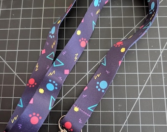 May include: A dark purple fabric lanyard featuring a repeating pattern of light blue and pink paw prints, yellow stars, pink diamonds, teal triangles, and yellow lightning bolts, finished with a silver-toned metal swivel lobster claw clasp.