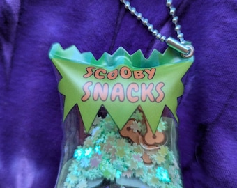 May include: A clear, plastic Scooby Snacks bag-shaped keychain with a green border and the words "Scooby Snacks" in orange. The bag is filled with glitter and a brown cartoon dog. A silver ball chain is attached to the top.