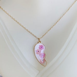 Op de afbeelding: Een gouden ketting met een roze en witte hartvormige hanger. De hanger is gemaakt van hars en heeft een glinsterende afwerking. Het hart is in twee&euml;n gebroken, met een helft die ontbreekt.