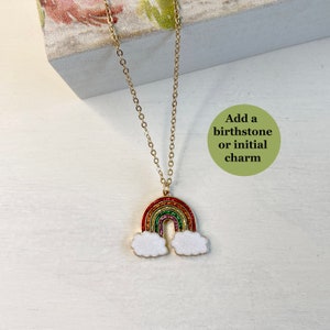 May include: A gold chain necklace with a rainbow charm and white cloud accents. The charm is covered in glitter. The text "Add a birthstone or initial charm" is written on a green circle.