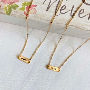 May include: Two gold necklaces with matching chain styles. The necklaces have rectangular pendants with the words "MAMA" and "MINI" engraved on them.