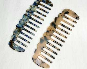 Può includere: Due pettini a denti larghi con bordo ondulato. Uno ha un motivo marmorizzato blu, marrone e bianco. L'altro ha un motivo tartarugato beige, marrone e nero. Entrambi i pettini sono su una superficie bianca.