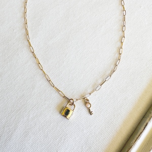 Op de afbeelding: Een gouden ketting met een klein hangslot en een sleutelbedel. De bedels hebben een subtiele glans.