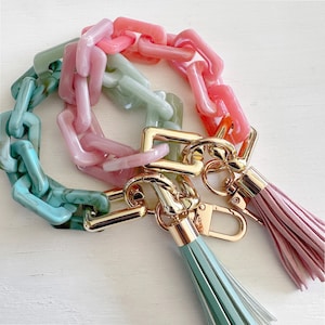May include: A multi-colored plastic chain with a gold-tone key ring and two tassels. The chain is made of pink, green, and white plastic links. The tassels are green and pink.