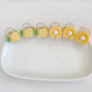 May include: A set of six small hair clips with yellow pineapple and daisy designs. The clips are clear plastic with a spring mechanism.