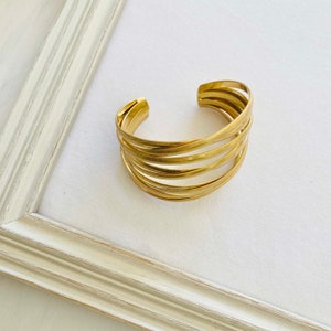 Op de afbeelding: Een gouden metalen armband met een gehamerde afwerking. De armband is gemaakt van meerdere dunne banden die op elkaar zijn gestapeld.