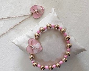 May include: A pink and gold necklace and bracelet set with a ballerina charm. The necklace is a delicate chain with a heart-shaped pendant featuring a ballerina in a pink tutu. The bracelet is made of gold beads with a pink heart-shaped charm featuring a ballerina in a pink tutu.