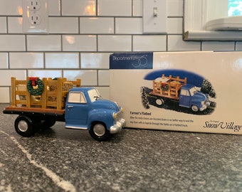 Puede incluir: Una figura de camión de plataforma azul con una caja de carga estilo madera, que presenta una pequeña corona verde con un lazo rojo. El camión se muestra junto a su caja original de Department 56 Snow Village, con el nombre Farmer's Flatbed.