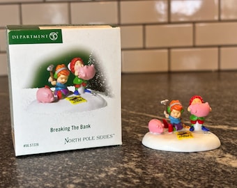 May include: A North Pole Series Christmas village figurine set depicting two children breaking a piggy bank. The figurine is titled "Breaking The Bank" and is part of the North Pole Series.