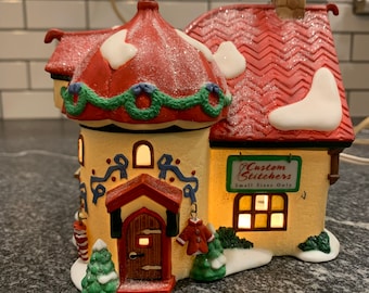 Op de afbeelding: Een kerstdorp huis van keramiek met een rood dak en witte sneeuw. Het huis heeft een bord met de tekst "Custom Stitchers Small Sizes Only".