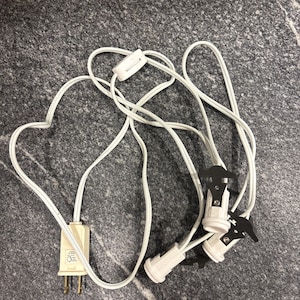 May include: White electrical cord with a plug, switch, and three light bulb sockets. The cord is coiled and arranged on a grey speckled surface. The plug has two flat prongs and a third round prong.