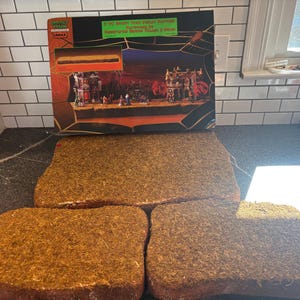May include: A Lemax Spooky Town 2-pc display platform set featuring a rectangular box with a graphic of a Halloween village scene and two separate, rectangular, brown, textured, grass-like foam display bases for miniature village setups.