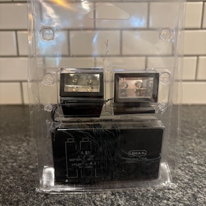 May include: A clear plastic package containing two small, rectangular light fixtures and a black battery pack. The light fixtures have a metallic appearance and are positioned above the battery pack. The battery pack has text that reads "1.5V" and "3 PILES A-4.5V".