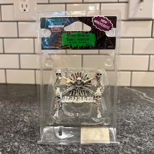 Puede incluir: Un banco de huesos en miniatura de color plateado con un diseño de calavera y huesos cruzados, empaquetado en un recipiente de plástico transparente. El banco tiene cuatro figuras de esqueleto y una calavera central. El embalaje incluye el texto "BONE BENCH" y "NEW NOUVEAUTÉ".