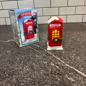May include: A red porcelain telephone booth figurine with white snow on the roof, a green wreath on the door, and yellow window panes. The item is accompanied by its original blue and white cardboard box labeled Dickensville Collectibles Noma.