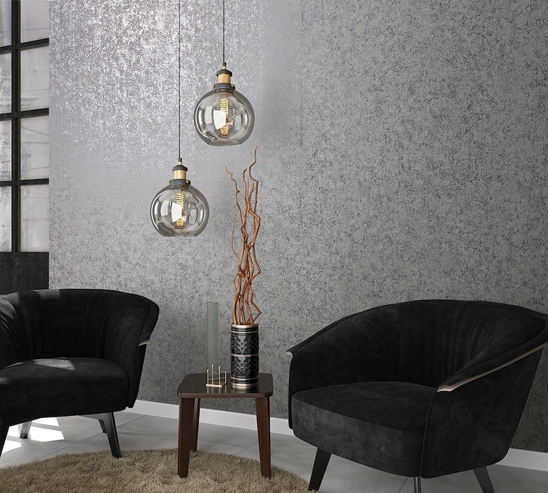 Op de afbeelding: Twee zwarte fauteuils met gouden accenten staan in een kamer met een getextureerde grijze muur. Twee glazen globe-hanglampen hangen aan het plafond. Een kleine houten tafel met een vaas met gedroogde takken staat tussen de fauteuils.