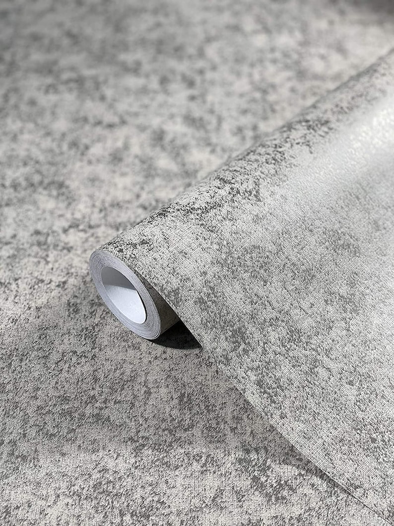 Op de afbeelding: Een rol grijze, gestructureerde behang met een subtiel gespikkeld patroon. Het behang heeft een licht ruwe, bijna betonachtige afwerking.