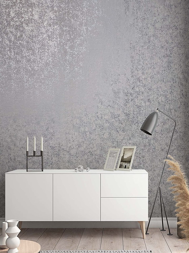 Op de afbeelding: Een witte kast met twee deuren en twee lades staat op een houten vloer voor een grijze, getextureerde muur. Een zwarte kaarsenhouder met drie kaarsen staat op de kast. Twee ingelijste foto's en een paar zilveren ornamenten staan ook op de kast. Een zwarte staande lamp met een witte verenpluim staat in de hoek van de kamer.
