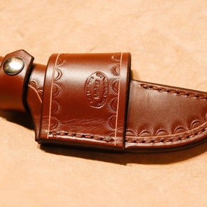 May include: A brown leather knife sheath with a decorative embossed pattern and a snap closure. The sheath is made by "Maker"