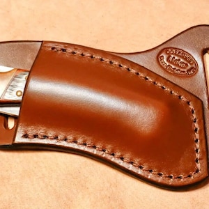 May include: A brown leather knife sheath with a stitched edge and a belt loop. The sheath has a maker's mark that reads "J.V. Leather, Made in the USA".