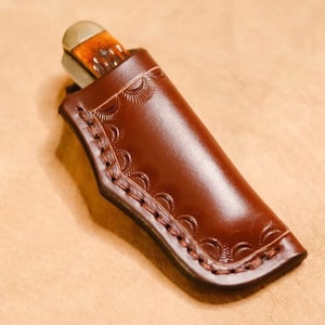 May include: A brown leather knife sheath with a decorative stitch pattern and a flap to secure the knife.