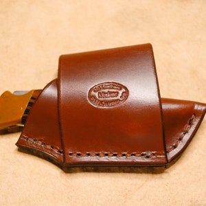 Könnte beinhalten: Eine braune Ledertasche für ein Messer mit einem genähten Rand und einem Herstellerzeichen mit der Aufschrift "Go Leather Maker, USA".