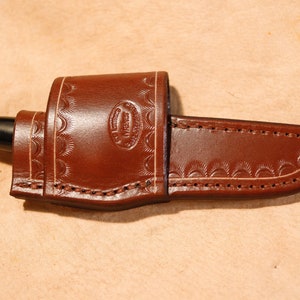 Può includere: Un fodero per coltello in pelle marrone con un motivo decorativo in rilievo e cuciture. Il fodero contiene un coltello con manico nero e lama argentata. Il fodero ha un logo stampato.
