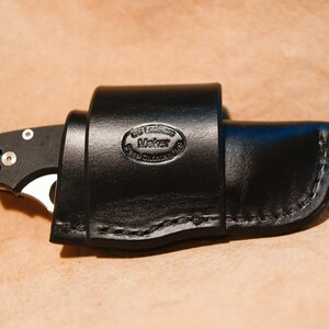 May include: A black leather knife sheath with a stitched edge and a maker's mark that reads "Leather Maker".