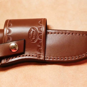 May include: A brown leather knife sheath with a snap closure and decorative stitching. The sheath has a maker's mark that reads "Leatherworks".