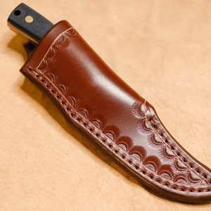 May include: A brown leather knife sheath with a decorative stitch pattern and a curved shape.