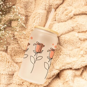 May include: A frosted glass tumbler with a wooden lid and a straw. The tumbler has a pink and white rose design.