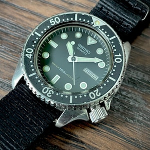 Könnte beinhalten: Eine Seiko Quartz Armbanduhr mit schwarzem Zifferblatt, leuchtenden Stundenmarkierungen und einer drehbaren schwarzen Lünette mit weißen Ziffern. Die Uhr verfügt über eine Datumsanzeige auf der Drei-Uhr-Position, silberne Zeiger und ein schwarzes Nylonarmband.
